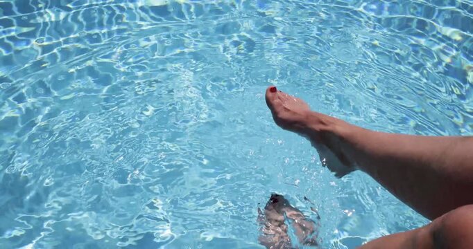 Slow motion footage of a pair of woman&rsquo;s feet gently splashing in a clear blue swimming pool with shimmering water reflections creating relaxing holiday scenes at a luxury resort during the summer