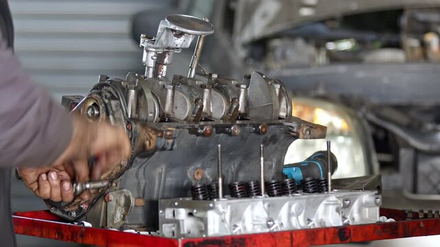 Mechanic hands working on a disassembled internal combustion engine block and cylinder head on a workbench in an auto repair shop. Metal automotive parts and mechanical repair tools with a vehicle blu