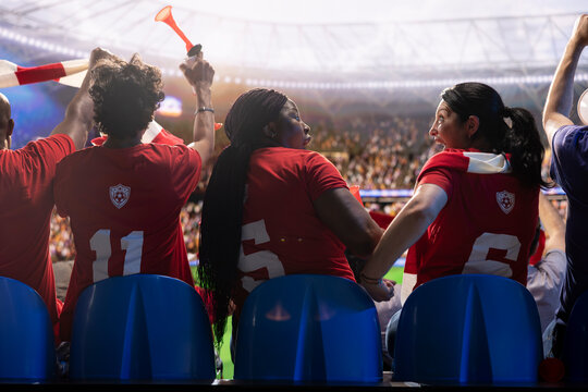 Female soccer supporters in red team shirts excited by goals scored during match, shocked women in stadium stands feeling thrilled by game moment on pitch, for sports fan culture and match day energy.