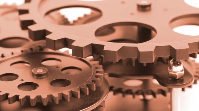 Close up view of metallic mechanical industrial gear wheels connected together. Orange toned machinery system parts, cogs, bolts, and interlocking components over a bright background. Technical engine