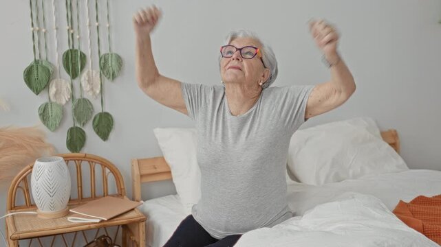 Elderly woman with glasses stretching both arms while sitting on bed in a residential building, bedside table and decor visible; morning routine serenity vitality.