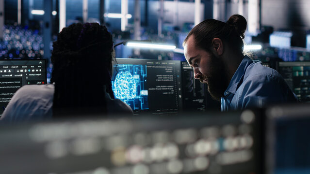 Server facility admin teaching trainee to check AI network working with AI. IT expert and African American woman at work applying AI to supervise servers powering ML.