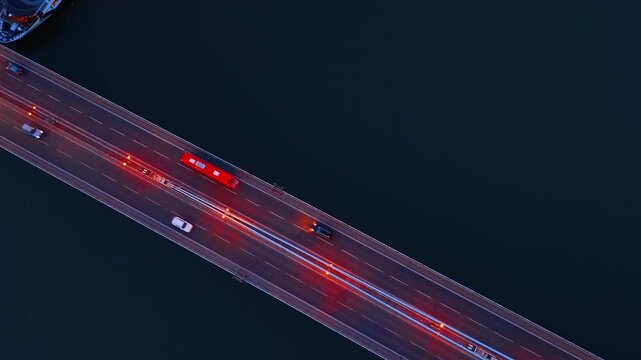 Aerial top down view of SNP Bridge traffic in Bratislava. A red bus and white cars drive on a illuminated asphalt road over dark water at night.