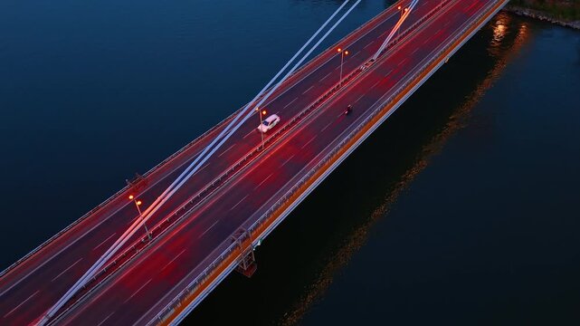 Low angle aerial view of SNP Bridge steel pylons. Long white cables stretch down to an illuminated roadway crossing dark water at twilight in Slovakia.