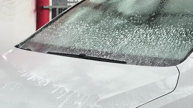 Thick layer of soap foam slides down vehicle windshield and hood at cleaning station. Self service carwash, pressure washer application, automotive body care.
