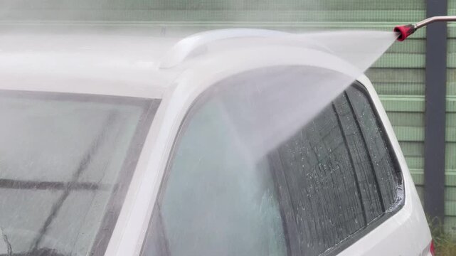 High pressure water jet sprays roof and windows of vehicle during cleaning process. Automotive maintenance, water resource management, consumer services.