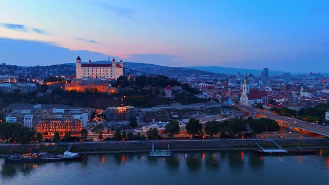 Bratislava Castle and old town skyline at twilight. Panoramic aerial view of the cityscape with illuminated cathedral and fortress under dusk sky.