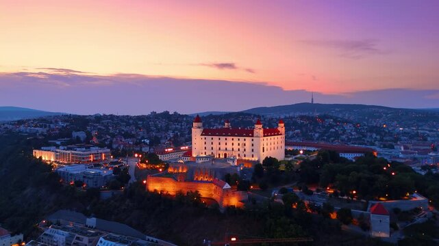 Bratislava Castle and city skyline at twilight in Slovakia. Distance view of illuminated historical fortress on hill surrounded by urban buildings at dusk.