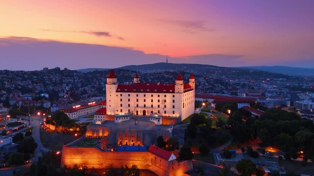 Illuminated Bratislava Castle at twilight in Slovakia. Close aerial view of white fortress building with red roofs during evening dusk.