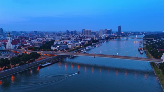 Danube River in Bratislava at dusk with a moving boat. Aerial view of the city bridges and illuminated skyline with water reflections during blue hour.