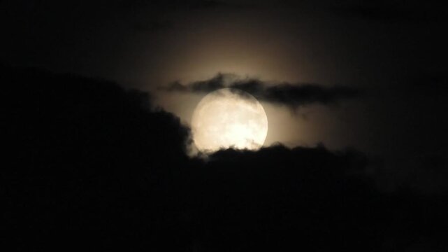 A full moon peeks out from behind clouds in a dark night sky. A close-up shot of the supermoon through a telephoto lens
