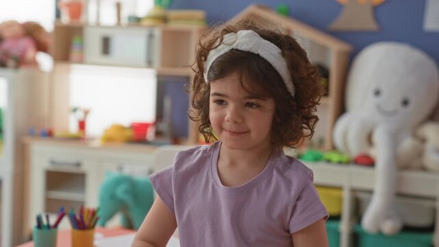 Little hispanic girl standing at a craft table with colored pencils, paper and plush octopus, smiling while turning her head and playing in building; childhood joy.