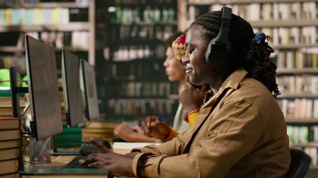 Woman student wearing headphones while working at desktop in library, black person surrounded by books, green lamps and study materials for digital learning, supporting education technology.