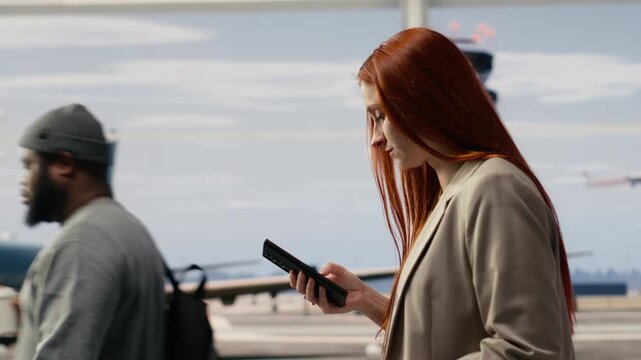 Close up of red haired woman walking in airport, holding phone and boarding pass, tourist holding plane ticket moving through airport terminal, using a smartphone to chat with friends.