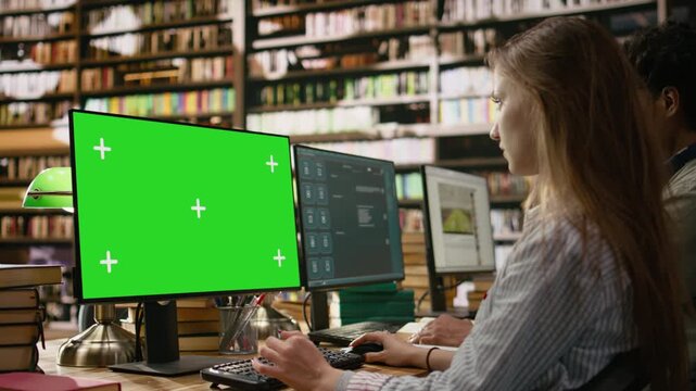Isolated green screen on desktop monitor with tracking markers on wooden library study desk, peers studying beside books, keyboards and bookshelves, computer class, for education technology.
