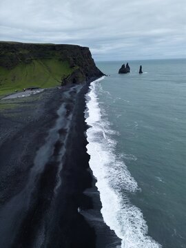 Aerial view of Reynisfjara Beach featuring its iconic black sand shoreline, dramatic sea stacks rising from the ocean, and lush green cliffs in Myrdalshreppur, Iceland.