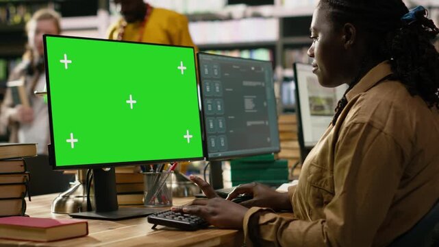 Green screen mockup on desktop with tracking markers shown near AI chatbot on monitor, student typing at library computer desk with books and shelves nearby, for editable screen mockup and VFX use.