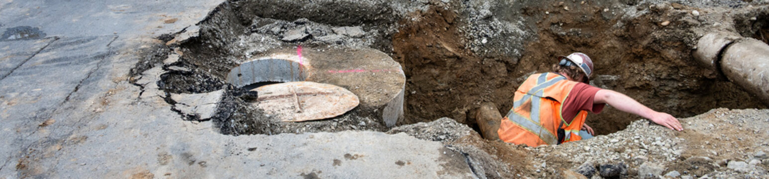 Underground utilities construction project, precast concrete stormwater system access vault in dirt trench under road construction worker in trench guiding excavator around old concrete pipe
