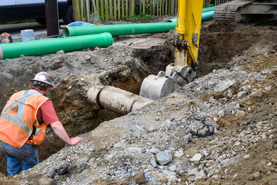 Excavator digging a deep narrow ditch in the dirt to work on underground utilities, construction worker in trench guiding machine operator around old concrete pipe
