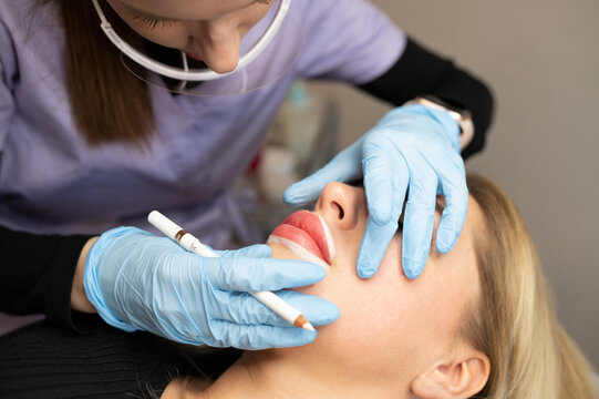Professional beautician drawing lips contour outline with a white cosmetic pencil before permanent makeup procedure