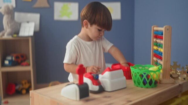 Boy playing with a toy cash register and wooden abacus, hands manipulating plastic pieces on a low table in a preschool classroom building with wall art and shelf toys; concentration.