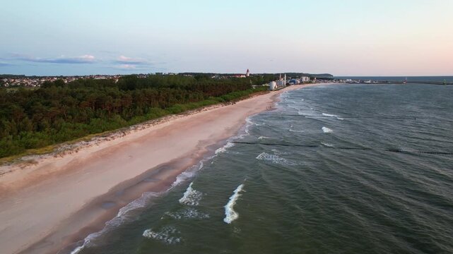 Scenic drone view flying over the Baltic sea coast, revealing a wide golden sand beach, dense green pine forest, and a distant coastal town during a peaceful summer dawn in Hel Peninsula, Poland.