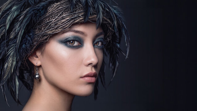 A glamorous woman with dramatic dark makeup wears a woven feathered headdress against a dark background, leaving ample copy space on the right side.