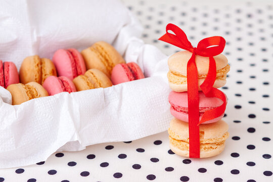 Three pink and beige macarons tied with a vibrant red ribbon beside an open white box filled with French sweets.