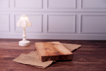 Soft glow warms wooden table's surface. Minimalist lamp stands beside rustic wood plank. Jute cloth adds texture beneath the wood. White wall paneling frames the tranquil scene © Nurma