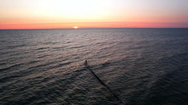 Vertical ascending aerial shot revealing a vast Baltic sea panorama during dawn. Drone flying upwards over wooden breakwater structures and calm waves in Hel Peninsula, Poland.