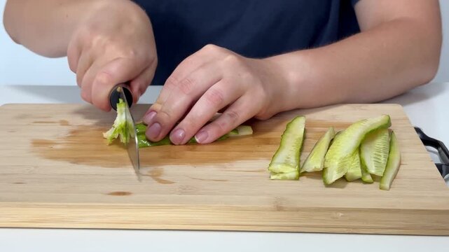 Woman using knife to thinly slice cucumber on cutting board in kitchen, preparing meal