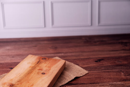 Warm wooden plank rests on burlap atop rich hardwood floor. Soft lighting highlights natural grain and subtle knots. Minimalist setup suggests calm, rustic kitchen or dining space