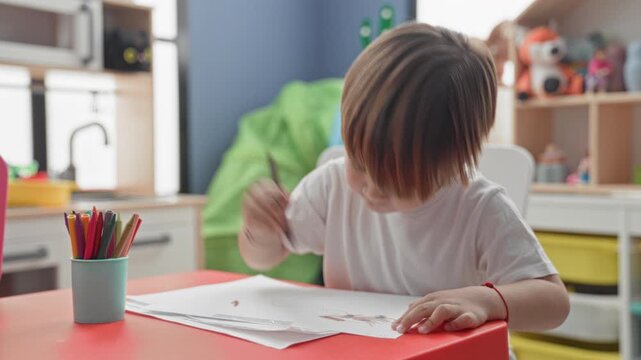 Boy holding drawing and pencil at red table in playroom with colored pencil cup, scattered papers and toy shelf visible; concentration creativity.