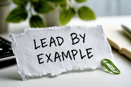 Torn paper with handwritten text Lead By Example on an office desk