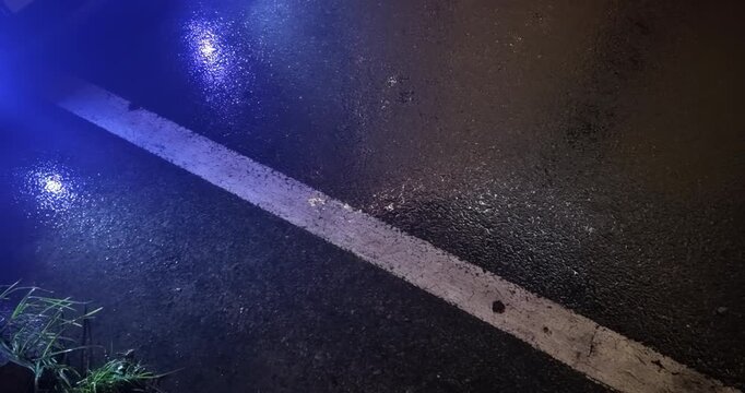 At night, under rain, the blue headlights of a parked car cast reflections onto the wet asphalt and the white lane marking.