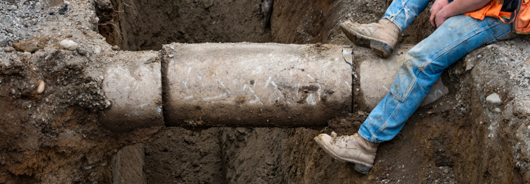 Construction worker sitting on edge of deep dirt trench by an exposed old concrete pipe, underground utilities municipal construction project
