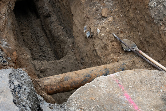 Deep narrow dirt trench with shovel and tamper bar, old iron pipe exposed in underground utilities construction project
