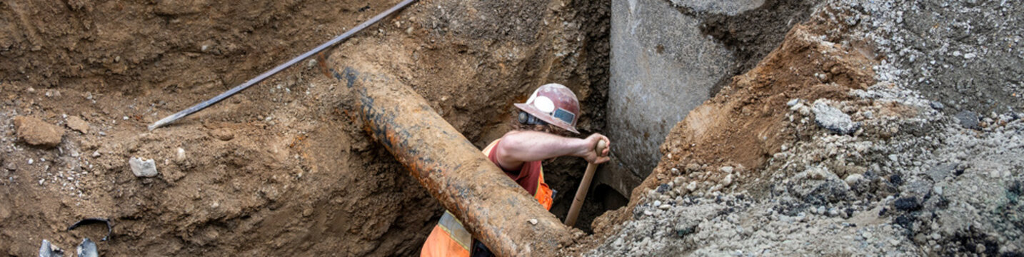 Construction worker in a deep trench digging dirt with a shovel around a precast concrete stormwater system access vault, underground utilities municipal infrastructure project

