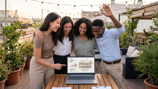 Diverse team of four young professionals cheering over laptop on creative co-working rooftop terrace
