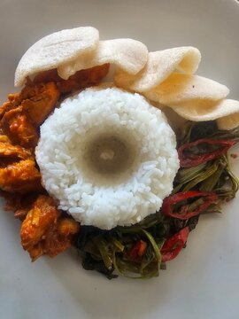 Top view of Indonesian lunch set featuring spicy chicken, plecing kangkung water spinach, ring-shaped white rice, and crackers on a white plate