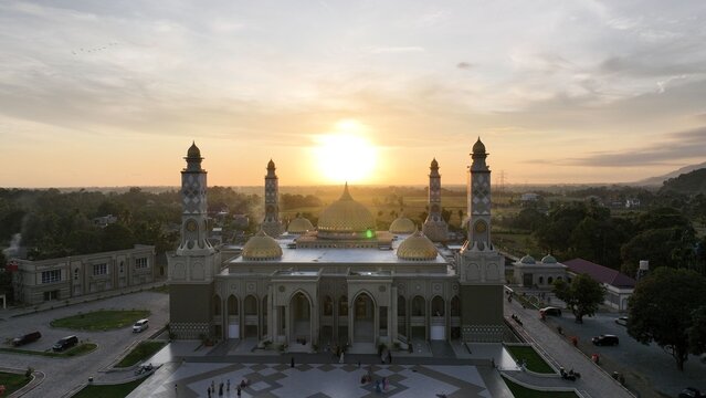 Aerial view of the Great Mosque of Blang Pidie at sunset, featuring golden domes, tall minarets, and a courtyard with people, surrounded by lush greenery in Blang Pidie, Aceh, Indonesia.