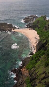 Aerial tropical beach cove with turquoise water and rocky headland waves breaking over coral reef, lush green vegetation cascading down cliffs, Bukit Merese, Lombok,Tanjung Ann, Indonesia