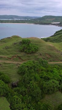 Group on cliff overlooking ocean, three hikers pause on grassy headland, aerial view reveals rolling waves, surf lines, distant islands and sandy bay under moody clouds,Bukit Merese, Lombok, Indonesia