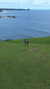 Couple Walking On Grassy Hill Overlooking Turquoise Ocean, Aerial Drone Shot Captures Sweeping Shoreline And Rolling Green Ridge. Two Travelers Stroll Along Winding Footpath Leaving Faint, Lombok