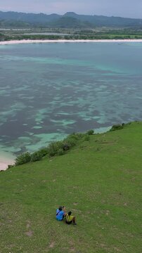 Couple sitting grassy hill overlooking bay, asian duo facing turquoise bay and coral reef, aerial hilltop view of vibrant shoreline, travel adventure vibe, shared quiet moment