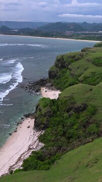 Aerial tropical beach cove with turquoise water and rocky headland waves breaking over coral reef, lush green vegetation cascading down cliffs, Bukit Merese, Lombok,Tanjung Ann, Indonesia