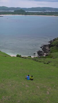 Couple sitting grassy hill overlooking bay, asian duo facing turquoise bay and coral reef, aerial hilltop view of vibrant shoreline, travel adventure vibe, shared quiet moment