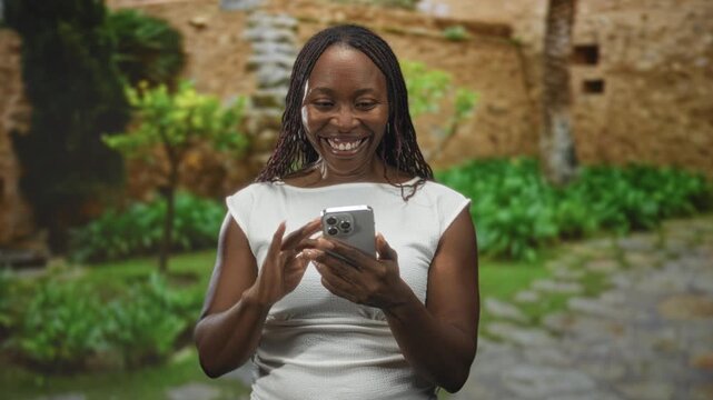 Young african american woman smiling while holding smartphone and tapping screen, hands visible, standing by stone wall building courtyard; joy connection.