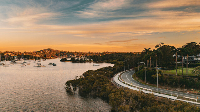 Aerial view of the coastal road winding along the waterfront with moored boats in the bay under a golden sunset sky in Bayview, New South Wales, Australia.