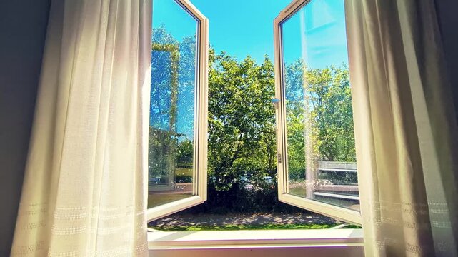Time lapse view from inside a room looking through an open white window. Lush green trees and a clear blue sky are visible outside on a bright sunny day, with moving shadows and a person walking by